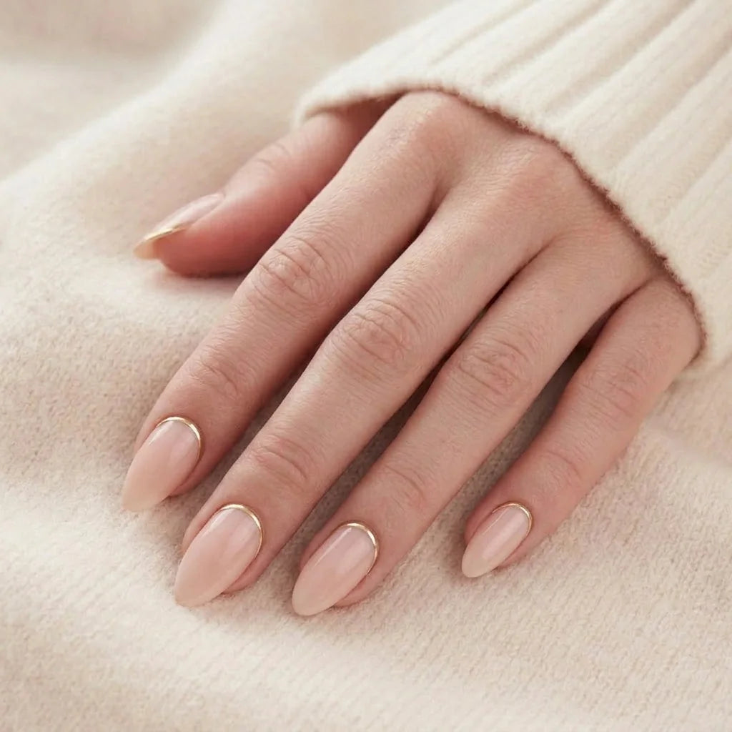 Golden Glimmer negler på hånd – peach nude mandel med gull-detalj mot beige strikket genser