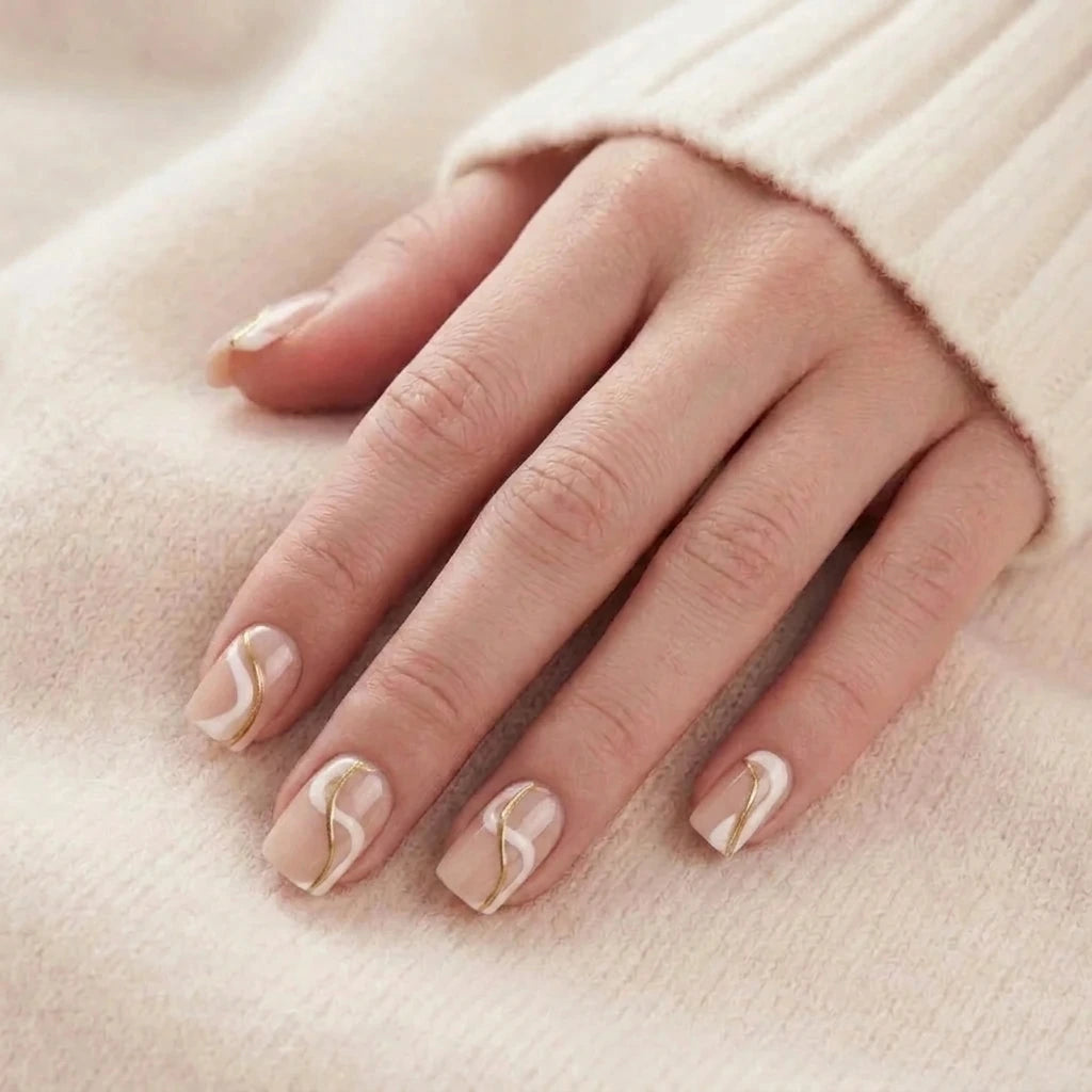 Korte, beige løsnegler med hvit og gyllen bølgedesign på en modellhånd mot en strikkegenser.